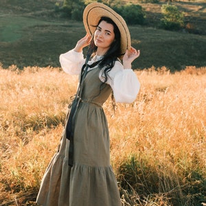 May include: A woman wearing a straw hat and a long, flowing, green dress stands in a field of tall grass.