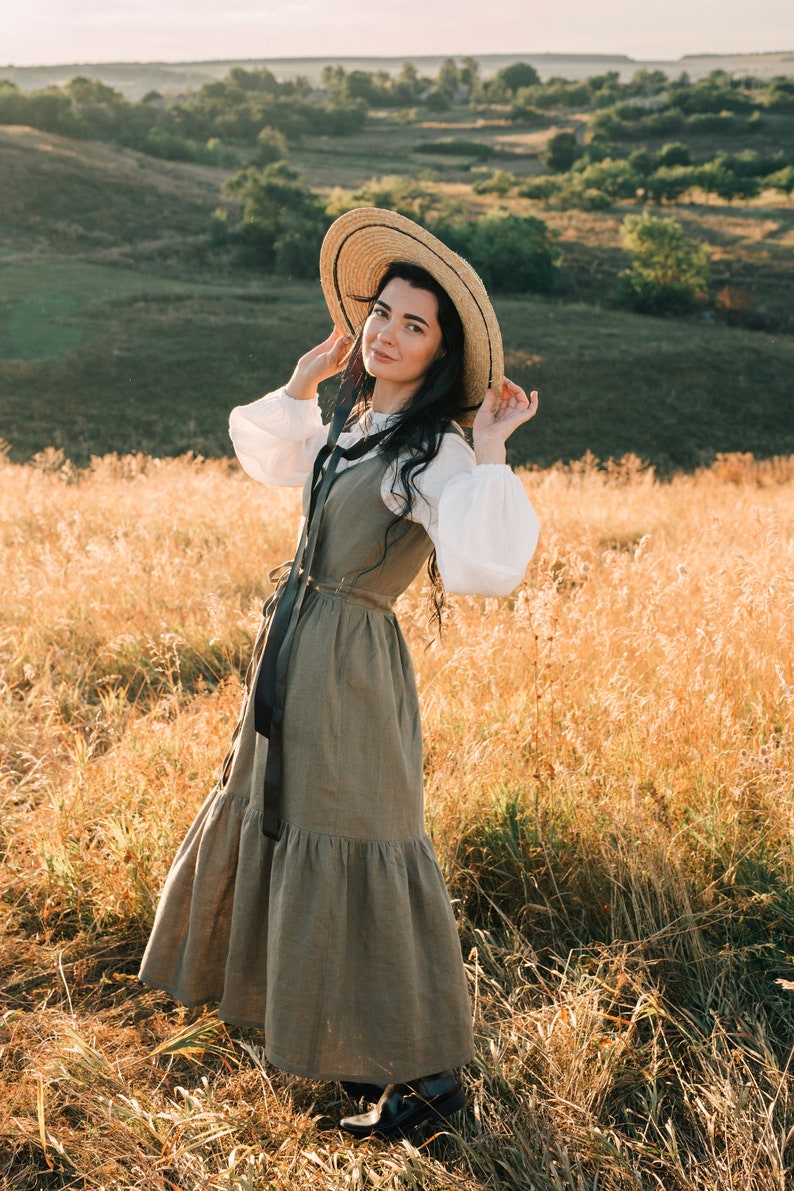 May include: A woman wearing a straw hat and a long, flowing, green dress stands in a field of tall grass.