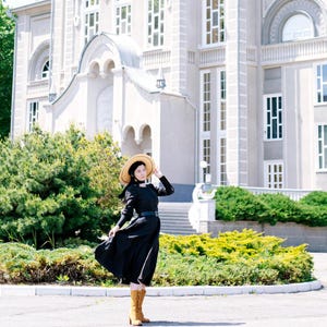 May include: A woman wearing a black dress and a straw hat stands in front of a white building. She is wearing brown boots and has her hair pulled back.