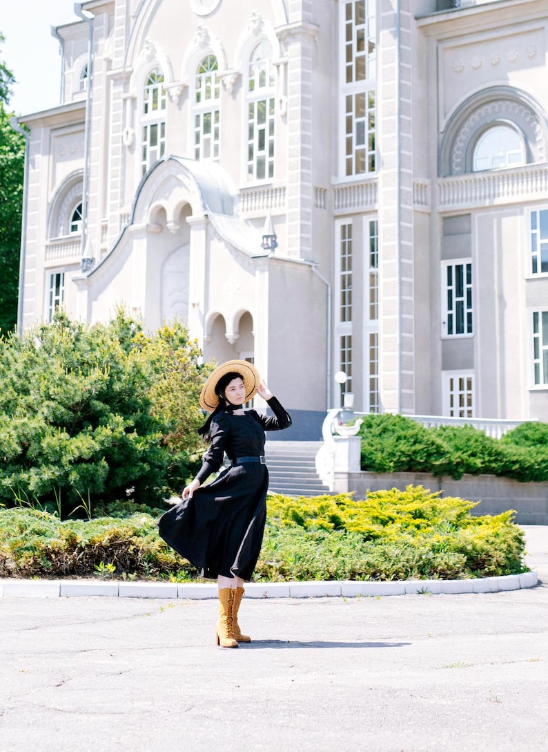May include: A woman wearing a black dress and a straw hat stands in front of a white building. She is wearing brown boots and has her hair pulled back.