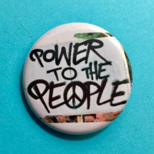 May include: A white button with a black and white image of a peace sign and the words "Power to the People" in black graffiti-style lettering.