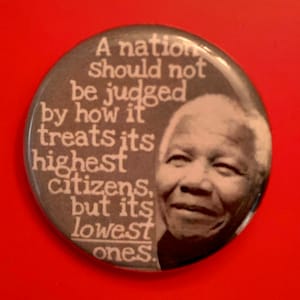 May include: A black and white button with a quote by Nelson Mandela: "A nation should not be judged by how it treats its highest citizens, but its lowest ones."