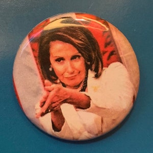 May include: A close-up of a button featuring a woman with a serious expression, wearing a white shirt and looking directly at the viewer. The background is a blurred image of an American flag.