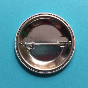 May include: The back of a silver button with a safety pin attached. The button has a white border around the edge.