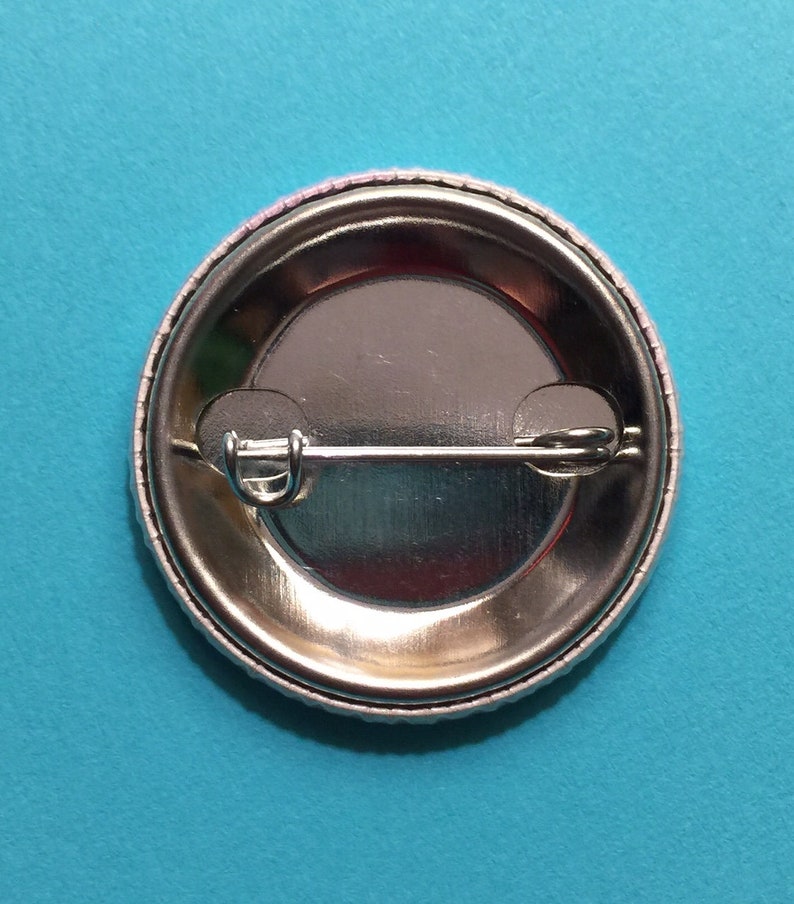 May include: The back of a silver button with a safety pin attached. The button has a white border around the edge.