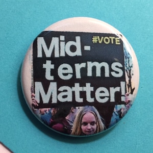 Può includere: Un bottone con un cartello in bianco e nero che dice "Mid-terms Matter!" con l'hashtag "#VOTE" in giallo. Il cartello è tenuto da un gruppo di persone, tra cui una giovane donna con i capelli lunghi e biondi.