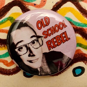 May include: A pink button featuring a black and white photograph of a person wearing glasses. The text "OLD SCHOOL REBEL" is printed in orange on the button.