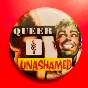 Könnte beinhalten: Ein runder Knopf mit gelbem Hintergrund und den Worten "Queer Unahamed" in roten Buchstaben. Der Knopf zeigt ein Cartoon-Bild eines Mannes mit einem lächelnden Gesicht.