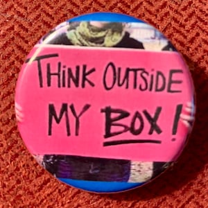 May include: A pink button with the text "Think Outside My Box!" written in black marker.