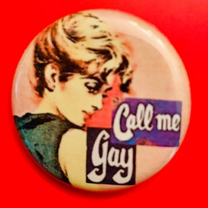 May include: A vintage-style button with a woman's face and the text "Call me Gay" in purple and white lettering on a pink, blue, and purple background.