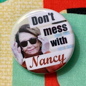 May include: A white button featuring a photograph of Nancy Pelosi wearing sunglasses and the text "Don't mess with Nancy".