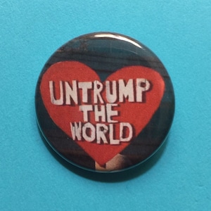 K&ouml;nnte beinhalten: Ein roter herzf&ouml;rmiger Knopf mit dem Text "Untrump the World" in wei&szlig;en Buchstaben.