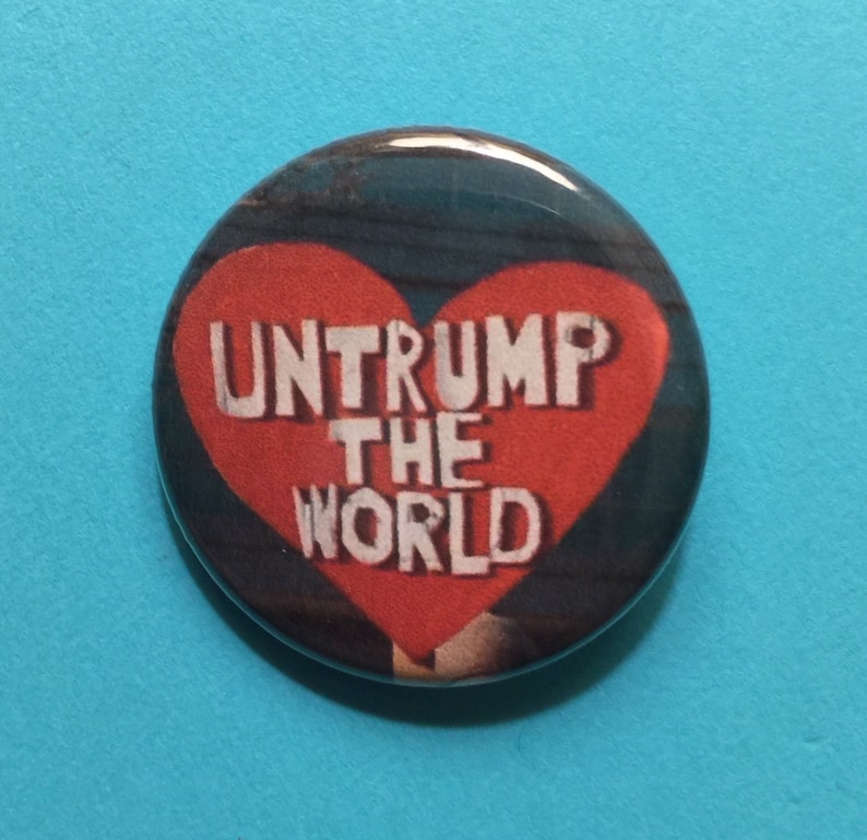 K&ouml;nnte beinhalten: Ein roter herzf&ouml;rmiger Knopf mit dem Text "Untrump the World" in wei&szlig;en Buchstaben.