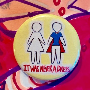 May include: A yellow button with a white outline of two figures, one wearing a blue shirt and red cape, the other in a white dress. The text "It was never a dress" is printed in red below the figures.