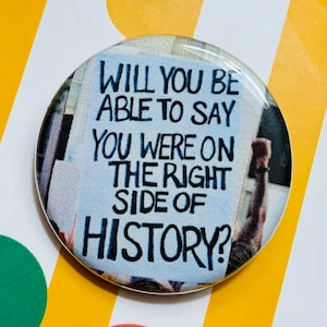 Peut inclure: Un bouton rond avec le texte "WILL YOU BE ABLE TO SAY YOU WERE ON THE RIGHT SIDE OF HISTORY?" en noir et bleu. Le bouton a une finition brillante et est placé sur un fond coloré.