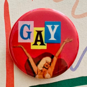May include: A red button with a white, blue, and yellow graphic that says "GAY". A woman with blonde hair is in the background with her arms raised.