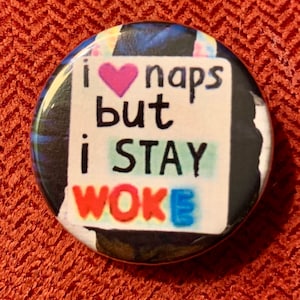 Puede incluir: Un botón redondo con un fondo colorido y un cuadrado blanco en el centro. El texto en el botón dice "I ♥ naps but I stay woke" con un corazón rosa.