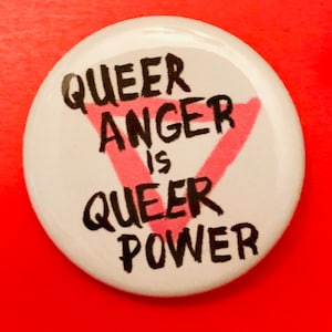 May include: White button with black text that reads "Queer Anger is Queer Power" with a pink upside down triangle in the center.