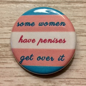May include: A blue, pink, and white button with the text "some women have penises get over it" printed on it.