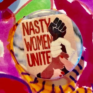 May include: A round button with a white background and red text that reads "Nasty Women Unite". A raised fist with a black hand and two white hands are shown on the button.