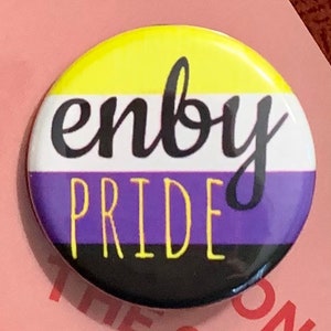 May include: A round button with the words "enby pride" in black and yellow lettering on a white, purple, black, and yellow background. The button is a symbol of non-binary pride.