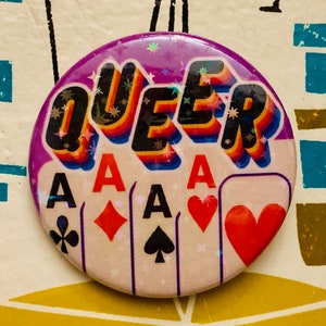 May include: A purple and white button with the word "QUEER" in rainbow letters and four playing cards with the ace of spades, diamonds, clubs, and hearts.