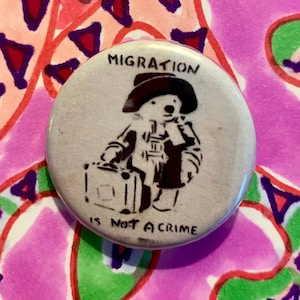 Può includere: Un bottone bianco con testo nero e un'illustrazione in bianco e nero di un orso con un cappello e una valigia. Il testo sul bottone dice "MIGRATION IS NOT A CRIME".