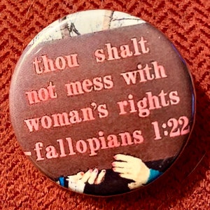 May include: A brown button with the text "Thou shalt not mess with woman's rights - fallopians 1:22" in pink lettering. The button features a photo of two hands shaking.