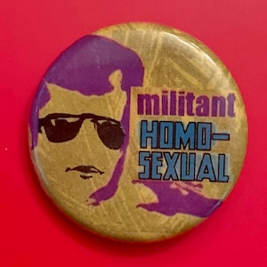 Könnte beinhalten: Ein runder Knopf mit gelbem Hintergrund und einer lila Silhouette einer Person mit Sonnenbrille. Der Text "militant HOMO-SEXUAL" ist in blauen und lila Buchstaben gedruckt.