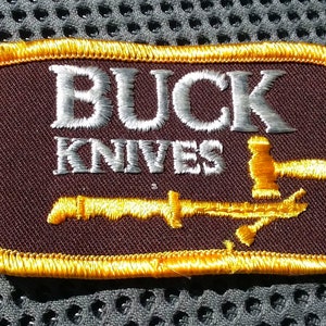 May include: A brown embroidered patch with a gold border. The patch features the text "BUCK KNIVES" in white embroidery and a stylized image of a knife and hammer in gold embroidery.