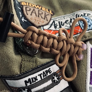 May include: A brown paracord knot tied around a green canvas bag with various patches, including "Bidwell Park Chico CA", "Adventure", "Mix Tape", and "Sacramento Kings".