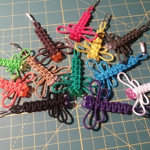 Könnte beinhalten: Eine Sammlung von 12 bunten Libellen-Schlüsselanhängern mit Perlenakzenten. Jeder Schlüsselanhänger ist aus geflochtener Schnur gefertigt und hat eine andere Farbkombination.
