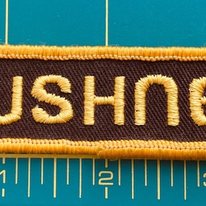 May include: A brown fabric patch with a yellow border and the word "BUSHNELL" embroidered in yellow.