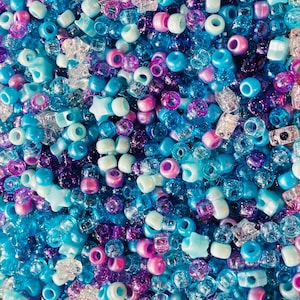 Op de afbeelding: Een close-up van een verzameling kleurrijke kralen in tinten blauw, paars, roze en helder. De kralen hebben verschillende vormen, waaronder rond, ster en buis. De kralen zijn verspreid, waardoor een textuurrijke en levendige weergave ontstaat.