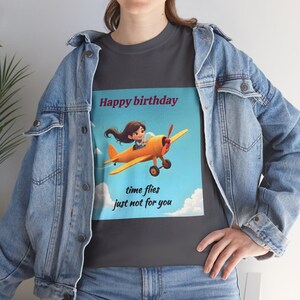 Può includere: T-shirt grigio scuro con un'illustrazione a cartoni animati di una ragazza su un aereo giallo contro un cielo blu con nuvole. Il testo "Happy birthday" è sopra l'immagine e "time flies just not for you" è sotto.
