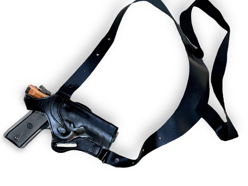 May include: Black leather shoulder holster with a handgun. The holster has a molded design to fit the gun and adjustable straps. The handgun is silver and black with a brown grip. The holster is designed to be worn under the arm.