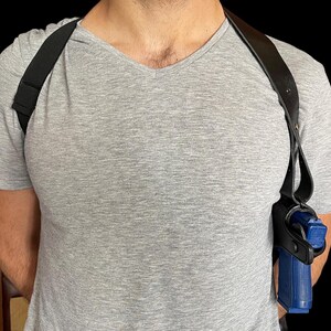 May include: Black leather shoulder holster with a blue handgun. The holster has adjustable straps and is designed to be worn under clothing. The image shows the holster on a person wearing a gray V-neck t-shirt.