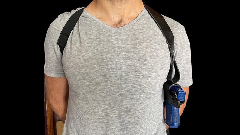 May include: Black leather shoulder holster with a blue handgun. The holster has adjustable straps and is designed to be worn under clothing. The image shows the holster on a person wearing a gray V-neck t-shirt.