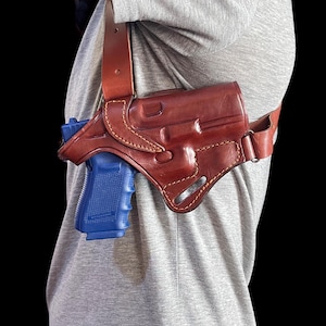 Könnte beinhalten: Ein braunes Schulterholster aus Leder mit einer blauen Schusswaffe. Das Holster ist mit hellbraunem Faden genäht und hat verstellbare Riemen. Die Schusswaffe ist im Holster sichtbar, mit dem blauen Griff und Lauf.