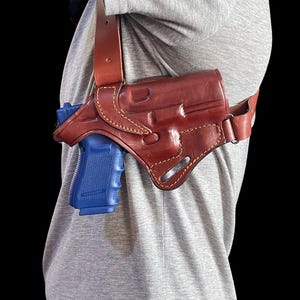 May include: A brown leather shoulder holster with a blue handgun. The holster is worn over a gray t-shirt. The leather is stitched with a lighter thread.