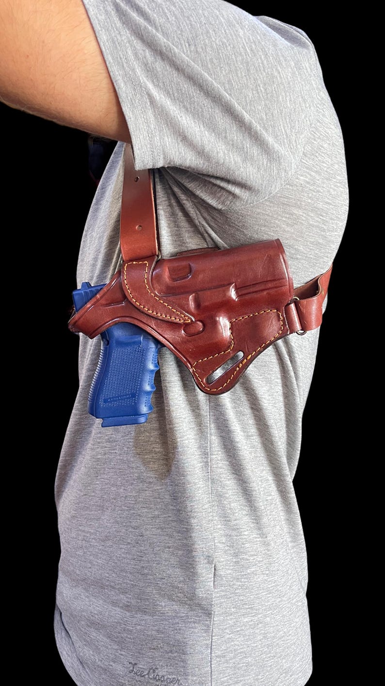 May include: A brown leather shoulder holster with a blue handgun. The holster is worn over a gray t-shirt. The leather is stitched with a lighter thread.
