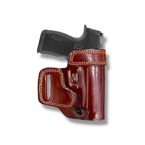May include: A black handgun with the text "SIG SAUER" is partially inserted into a brown leather holster. The holster has visible stitching and a black button closure. The holster is designed to be worn on the body.