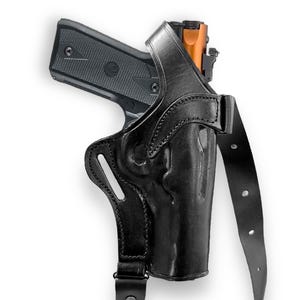 May include: Black leather shoulder holster with a semi-automatic pistol. The holster features a molded design and adjustable straps. The pistol has a black frame and an orange-colored slide. The image is set against a white background.