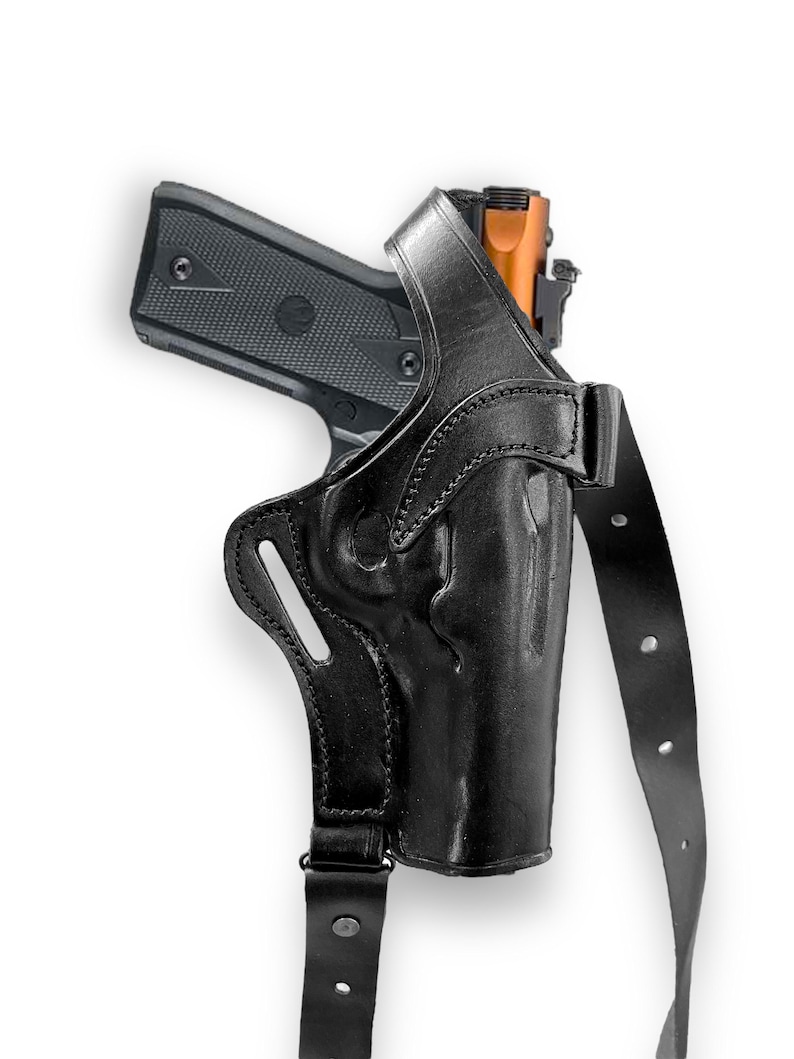 May include: Black leather shoulder holster with a semi-automatic pistol. The holster features a molded design and adjustable straps. The pistol has a black frame and an orange-colored slide. The image is set against a white background.