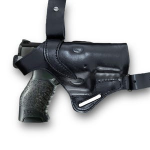 May include: Black leather shoulder holster with a semi-automatic pistol. The holster has a molded design to fit the gun and features adjustable straps for a secure fit. The pistol is black and gray with a textured grip.