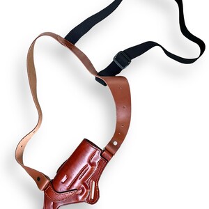 May include: A brown leather shoulder holster with a black adjustable strap. The holster is designed to hold a handgun and features detailed stitching and a molded shape. The leather has a rich, polished finish.