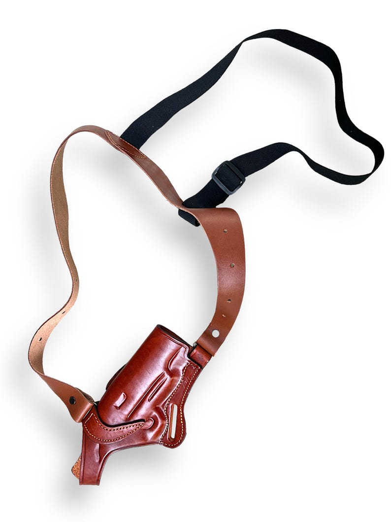 May include: A brown leather shoulder holster with a black adjustable strap. The holster is designed to hold a handgun and features detailed stitching and a molded shape. The leather has a rich, polished finish.