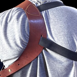 May include: A brown leather shoulder holster with a black elastic strap, worn over a gray t-shirt. The holster has a stitched design and brass-colored hardware. The background is black.