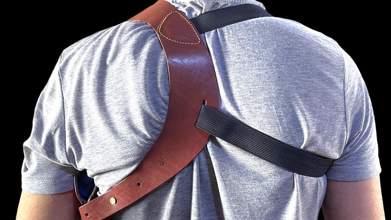 May include: A brown leather shoulder holster with a black elastic strap, worn over a gray t-shirt. The holster has a stitched design and brass-colored hardware. The background is black.