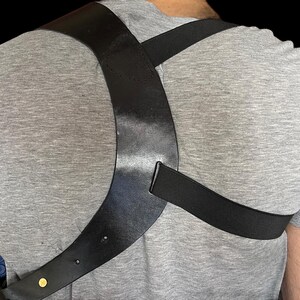 May include: Black leather shoulder holster with a cross-body design. The holster is worn over a gray t-shirt. The holster has a strap that goes over the shoulder and another that goes around the torso. A gold-colored rivet is visible.